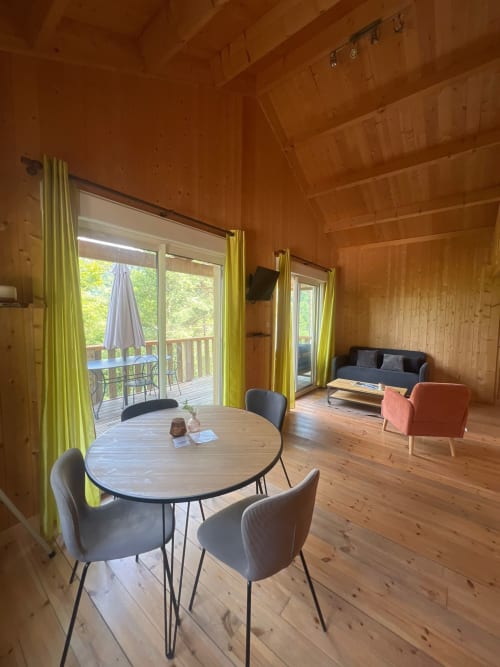 Interno di lodge in legno moderno con tavolo da pranzo, zona living e terrazza affacciata sul fiume presso Cabanes du Belvédère