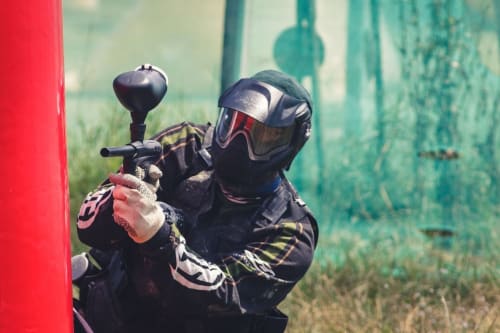 Jugador de paintball en equipo táctico apuntando marcador hacia pared roja, actividad de tradición rural en Nieva