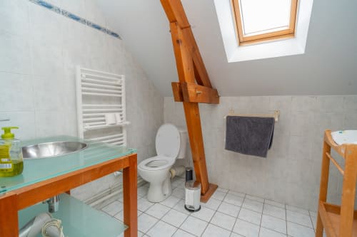 Salle de bain lumineuse sous les combles aux Vacances en Touraine avec poutres bois, toilettes blanches, lavabo turquoise et puits de lumière.
