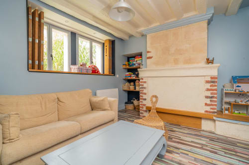 Lumineux salon de jeux à Vacances en Touraine avec canapé beige, cheminée, jouets et grandes fenêtres sur le jardin