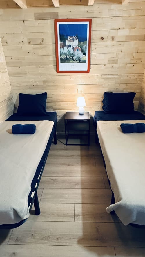 Camera matrimoniale con letti bianchi e biancheria blu navy in una rustica lodge in legno, comodino centrale con lampada, quadro di montagna