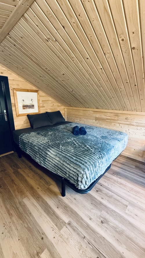 Accogliente camera loft nel lodge in legno con soffitto mansardato, biancheria blu e cuscini neri alle Cabanes et lodges du Belvédère
