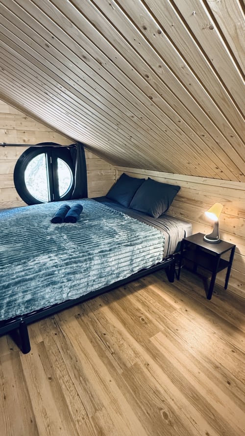 Accogliente camera da letto in mansarda al Lodge l'EPERVIER con finestra circolare, soffitto in legno, biancheria blu, comodino nero e lampada