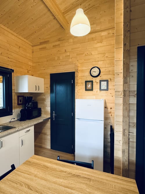 Cucina di lodge in legno moderno con elettrodomestici bianchi, porta nera, lampada a sospensione e orologio presso Cabanes et lodges du Belvédère