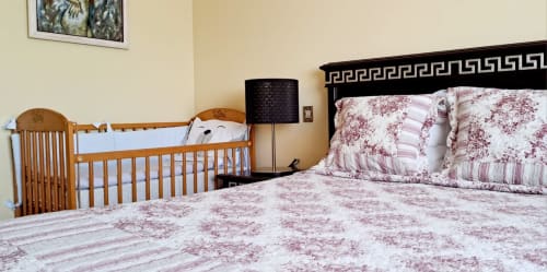 Quarto na Casa Da Avó Ilda com berço de madeira, cama queen com almofadas florais, luminária e paredes creme em Matosinhos.