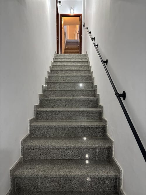 Escalera de granito en el edificio Holibai, Baiona, que conduce a puerta de madera con iluminación cálida y barandilla metálica.