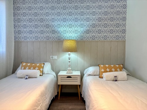Dormitorio doble en apartamento Holibai, Baiona. Dos camas con almohadas naranjas, mesita de noche de madera, lámpara amarilla, papel pintado estampado.