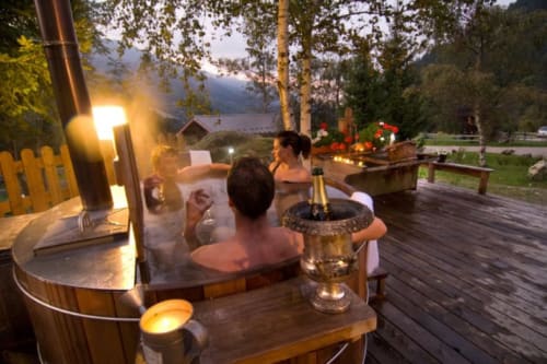 Guests relaxing in a wooden hot tub at Auberge Nemoz, with champagne and mountain lake views at dusk