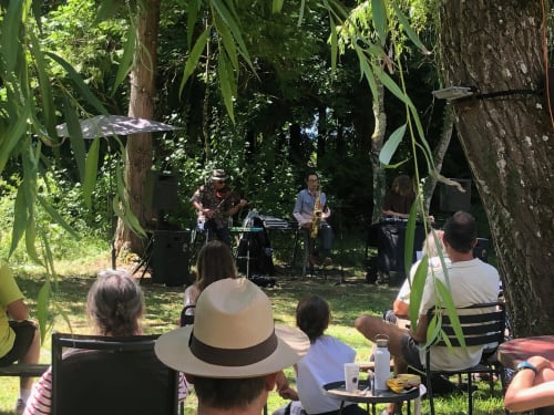 Spectacle de jazz en direct dans le jardin ombragé du Domaine de l'Ocrerie, Pourrain. Les invités écoutent la musique sous la canopée.
