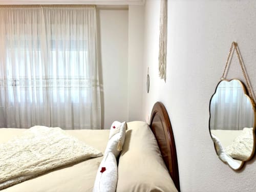 Luminoso dormitorio en apartamento de Baiona con cama, espejo decorativo, cortinas translúcidas y vistas al mar