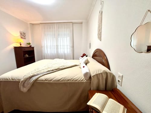 Dormitorio luminoso en apartamento de Baiona con cama de madera, ropa de cama crema, espejo y cortinas translúcidas con vistas al mar