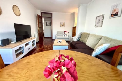 Luminoso salón del apartamento de Baiona con mesa de comedor de madera, flores rojas, televisor, sofá y vistas al mar