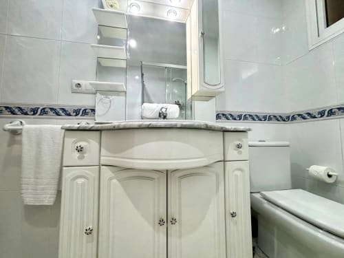 Baño moderno con tocador blanco, espejo iluminado y cenefa decorativa de azulejos azules en apartamento de Baiona