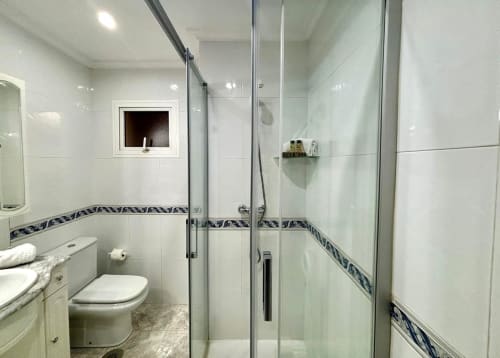 Baño moderno con ducha de cristal, paredes de azulejos blancos, cenefa decorativa azul, inodoro y lavamanos en Exeira
