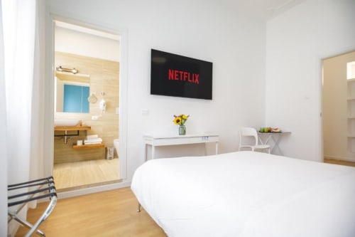 Modern bedroom at Bluemoon Sardinia in Cagliari with white bed, wall-mounted TV showing Netflix, desk, and ensuite bathroom