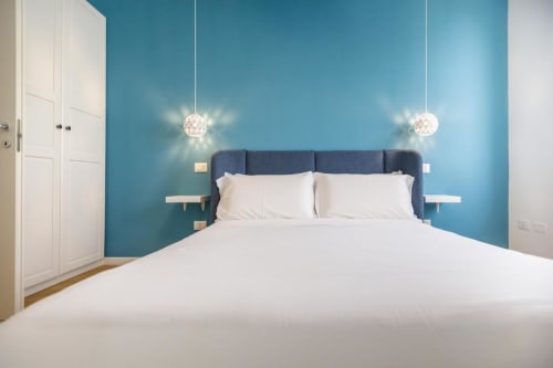Modern bedroom at Bluemoon Sardinia in Cagliari with blue accent wall, white bedding, pendant lights, and floating