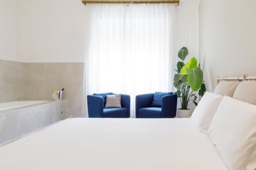 Modern bedroom at Bluemoon Sardinia in Cagliari with white bedding, blue armchairs, bathtub, and city views through sheer