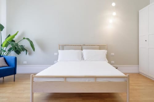 Modern bedroom at Bluemoon Sardinia in Cagliari with light wood bed, white bedding, pendant lights, and blue accent chair