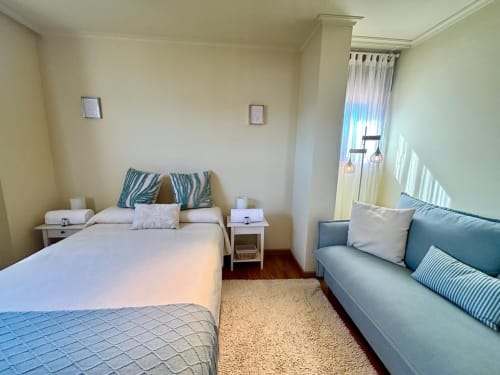 Dormitorio luminoso con cama, sofá azul y balcón con vista al mar en apartamento de Baiona