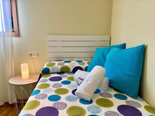Dormitorio luminoso con ropa de cama de lunares coloridos, almohadas turquesas, cabecero blanco y lámpara de noche en apartamento de Baiona