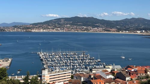 Bahía de Baiona con veleros y pueblo costero anidado en colinas verdes frente a las aguas azules del Atlántico