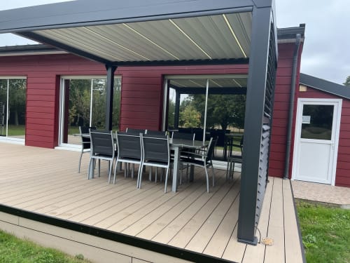 Terrasse moderne avec pergola rétractable, table à manger et maison rouge du gîte CafecouetBreizh à Guipry-Messac, France