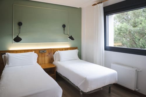Habitación doble en Hostal El Ciervo con sábanas blancas, cabecera de madera, pared verde salvia y gran ventana con vistas a los árboles