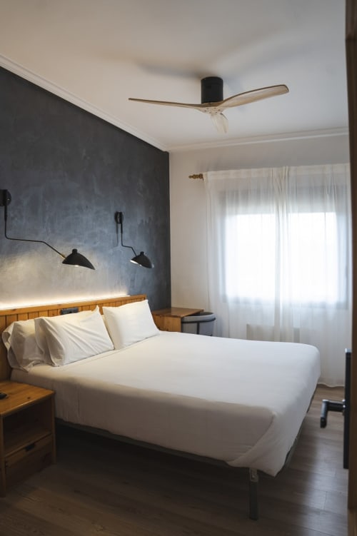 Habitación moderna en Hostal El Ciervo con cama blanca, cabecero de madera, pared de acento oscuro, ventilador de techo y luz natural