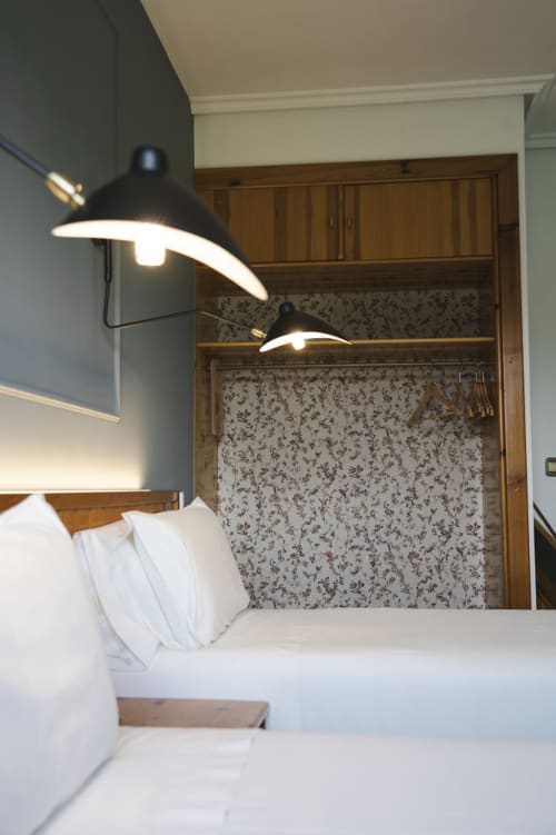 Habitación moderna en el hostel El Ciervo con cama de sábanas blancas, muebles de madera, papel tapiz floral y lámparas de lectura negras