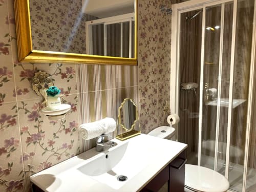 Baño moderno en apartamento Vermello, Baiona con espejo iluminado, lavabo blanco, ducha y papel tapiz floral