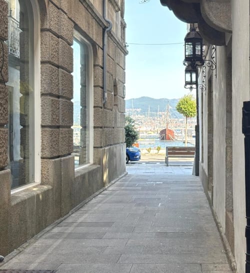 Estrecho callejón en Baiona con edificios de piedra que desembocan en vistas al puerto con botes y montañas