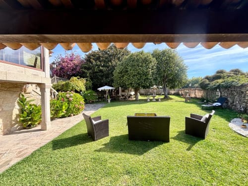 Jardín soleado de la villa Holibai en Baiona con zona de descanso, césped frondoso, árboles en flor y muros de piedra