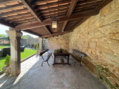 Terraza cubierta de piedra con techo de vigas de madera, mesa de comedor, sillas y vistas al jardín en la villa Miña Xoia de Baiona