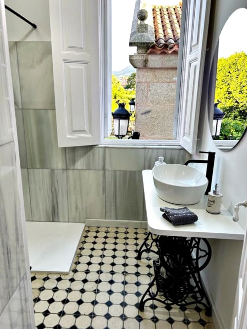 Baño elegante en villa Holibai con lavabo de pedestal, piso de azulejos blanco y negro, ventana abierta hacia el jardín de Baiona
