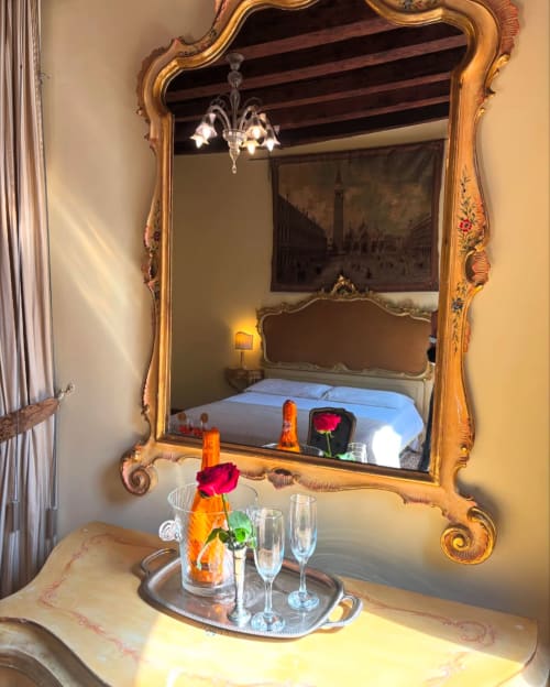 Specchio ornato con cornice dorata che riflette una camera da letto lussuosa con lenzuola blu nel palazzo veneziano, rose fresche e champagne su marmo