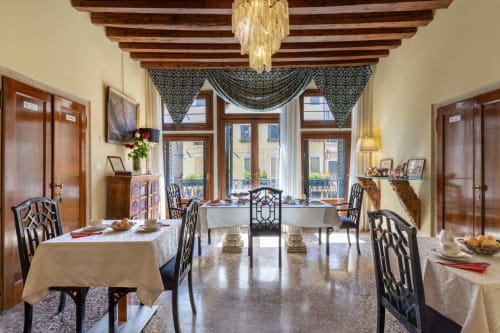 Elegante sala da pranzo veneziana con travi in legno a vista, tavoli formali con sedie decorate e vista sul mare