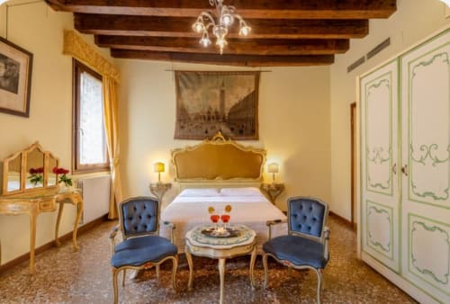 Camera da letto di palazzo veneziano lussuoso con testata dorata ornata, sedie blu, travi a vista e arredi eleganti a Venezia