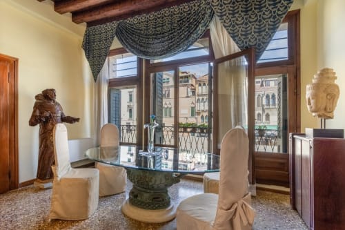 Suite elegante di palazzo veneziano con tavolo in vetro, sedie imbottite, balcone con vista sul canale e drappi ornamentali
