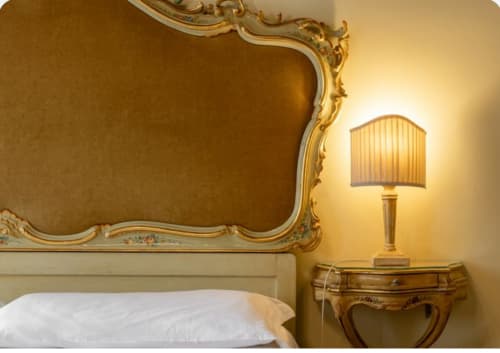 Camera da letto elegante presso Palazzo San Firmino Venezia con testiera dorata ornamentale, illuminazione calorosa e comodino antico