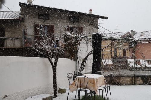 Cortile innevato dell'Appartamento Sormano 3 con alberi spogli, tavolo da pranzo e edifici in pietra