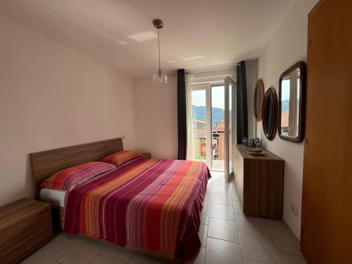 Camera luminosa nell'appartamento di Sormano con letto a righe rosse, vista montagna, arredi moderni e accesso al balcone