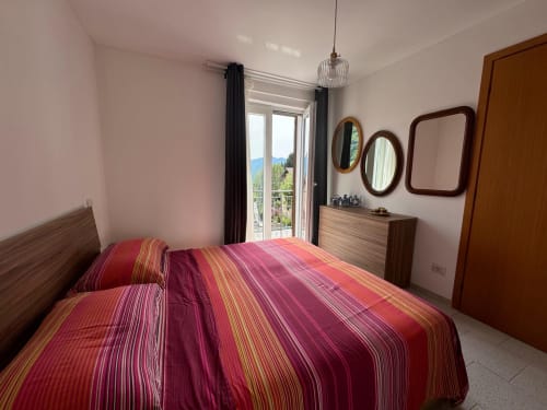 Camera da letto dell'Appartamento Sormano 2 con biancheria rossa rigata, vista montagna dal balcone e mobili in legno