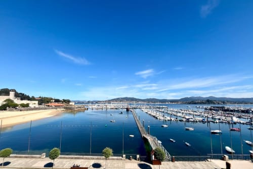Marina de Baiona con veleros, playa y edificios costeros bajo un cielo azul despejado