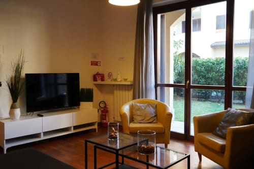 Moderno soggiorno dell'Appartamento Roma 2 con pareti gialle, poltroncine in pelle abbronzata, TV, porte con vista sul giardino e pavimenti in legno