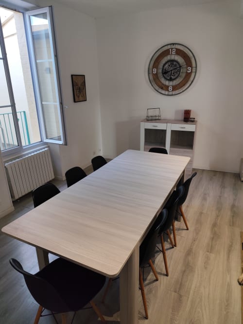 Salle à manger moderne de l'appartement Accolade à Châteaudun avec table en bois clair, chaises noires et vue sur la ville depuis la porte-fenêtre du balcon