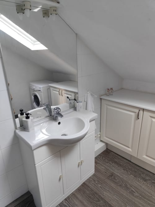 Salle de bain lumineuse sous les toits de l'appartement Accolade à Châteaudun avec lavabo blanc, miroir, lave-linge et plafond mansardé