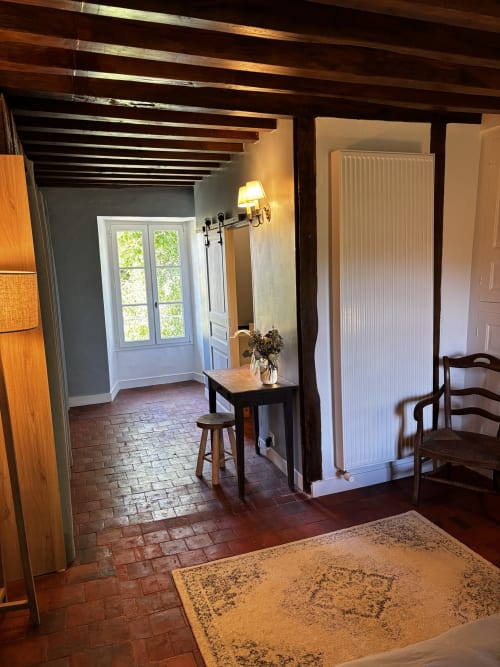 Chambre secrète au Prieuré Du Preux avec poutres apparentes, carrelage en terre cuite, portes blanches, vue sur jardin et charme rustique