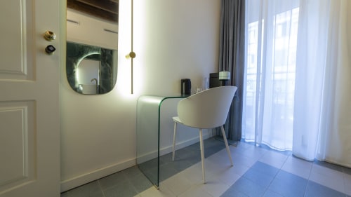 Modern bedroom vanity with illuminated mirror, curved glass desk, white chair, and city view at YOUnique Boutique Rooms