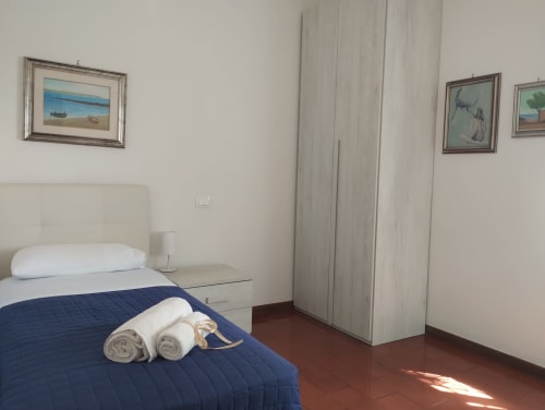 Camera luminosa con letto matrimoniale, coperta blu navy, asciugamani arrotolati, comodino con lampada, quadri costieri e armadio