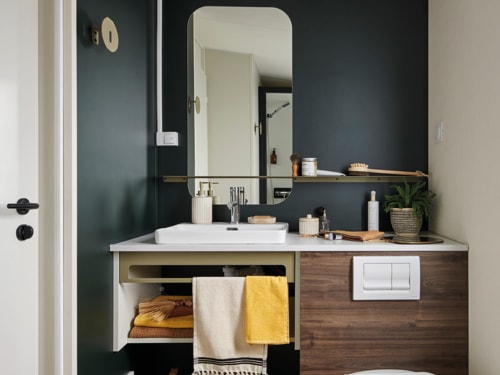 Salle de bain moderne de l'hôtel Le Maquis Plage avec murs bleu foncé, vasque blanche, meuble en bois et miroir à cadre doré